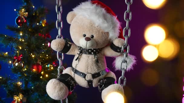 Ένα αρκουδάκι σε ένα καπέλο Άγιος Βασίλης είναι ένα χριστουγεννιάτικο δώρο για BDSM παιχνίδια στο φόντο ενός χριστουγεννιάτικου δέντρου — Αρχείο Βίντεο
