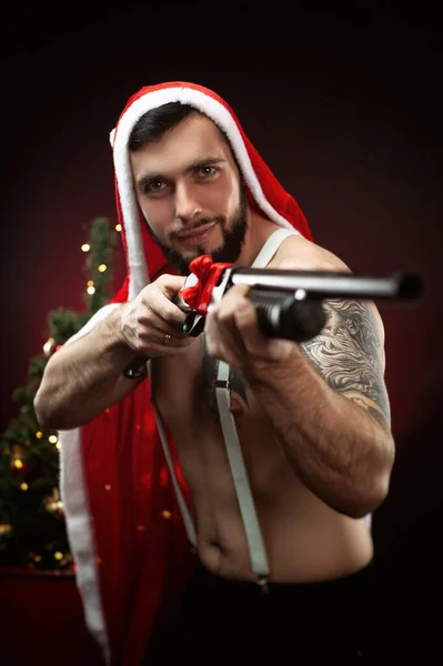 Silny facet w czerwonej pelerynie Świętego Mikołaja na choince ze strzelbą w ręku — Zdjęcie stockowe