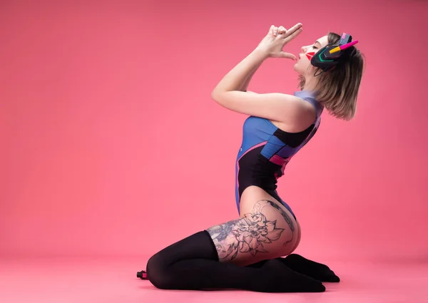 Gadis gamer seksi dalam mengungkapkan bodysuit cerah dan headset gaming pada latar belakang merah muda Stok Foto
