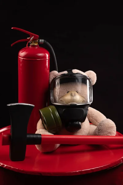 The mascot of the fire brigade is a teddy bear in a gas mask with a fire extinguisher and a red axe in smoke on a dark background — Stockfoto