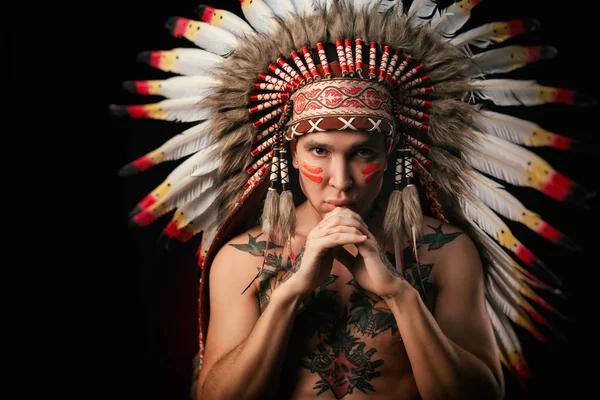 A white man in a headdress of the indigenous peoples of North America with a naked torso on a dark background — Stock Photo, Image