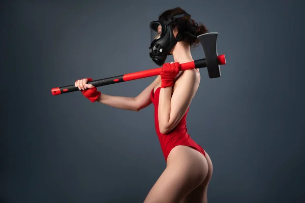 Flicka i en röd body i en gasmask med en brandyxa på en grå bakgrund — Stockfoto