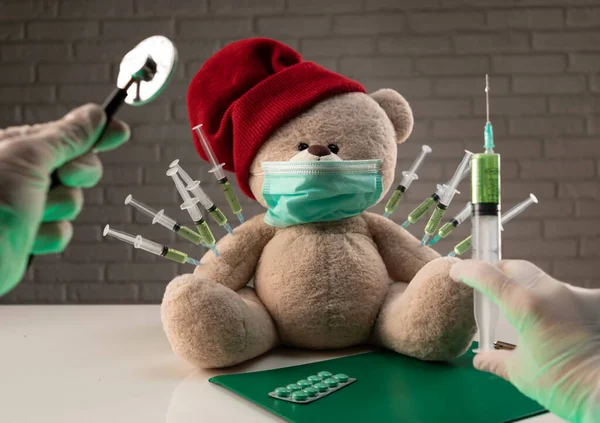 Pluszowy miś w masce medycznej ze strzykawkami w ramieniu na stole lekarskim otrzymuje drugie szczepienie przeciwko wirusowi korony — Zdjęcie stockowe