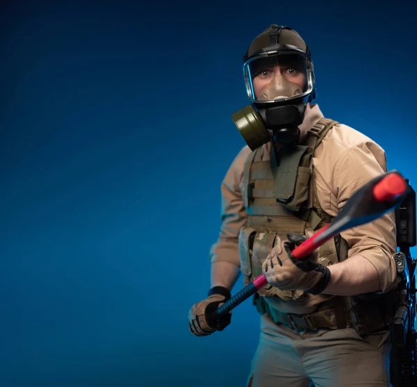 A soldier in military clothes with a bulletproof vest in a gas mask is holding a red fire axe in his hands in fright — Stock Photo, Image