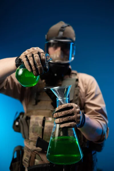 Un uomo in abiti militari e una maschera antigas con fiaschi chimici da laboratorio versa reagenti — Foto Stock