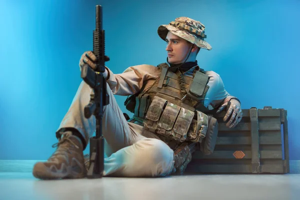 Ένας άντρας στρατιώτης με καμουφλάζ κάθεται σε ένα κιβώτιο πυρομαχικών στο πάτωμα με ένα όπλο. — Φωτογραφία Αρχείου