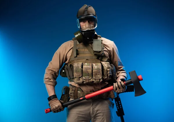 Voják ve vojenských šatech s neprůstřelnou vestou v plynové masce drží v rukou červenou sekeru — Stock fotografie