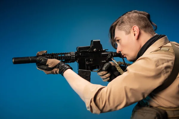 Ein männlicher Spezialagentensoldat in einer kugelsicheren Weste mit einem automatischen Gewehr auf blauem Hintergrund — Stockfoto