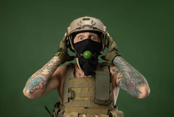 A soldier man in military clothes helmet with a bdsm gag in his mouth expresses emotions, photo joke — Stock Photo, Image