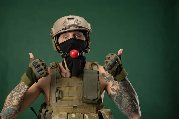 Un soldado con casco de ropa militar con una mordaza bdsm en la boca expresa emociones, foto broma —  Fotos de Stock