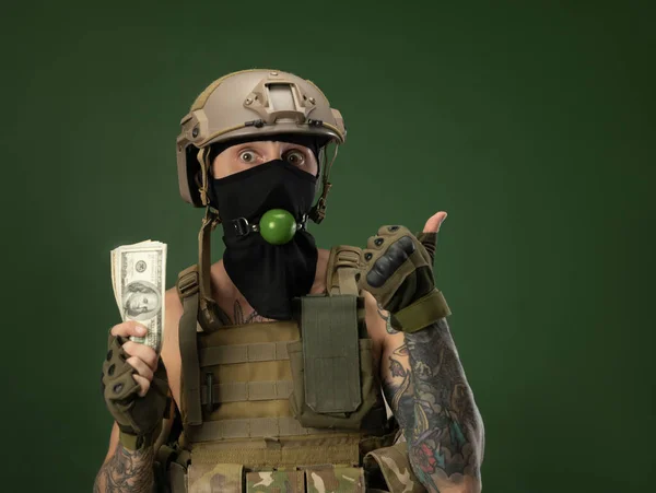 Um soldado homem em capacete roupas militares com uma mordaça bdsm em sua boca expressa emoções segurando dinheiro dólares em suas mãos, piada foto — Fotografia de Stock