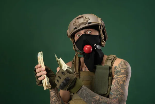 Żołnierz w wojskowym hełmie z kneblem bdsm w ustach wyraża emocje trzymając pieniądze w rękach, żart fotograficzny — Zdjęcie stockowe