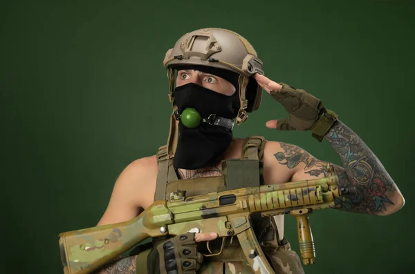 Un soldato di sesso maschile in abiti militari, un casco con un bdsm bavaglio in bocca esprime emozioni tenendo in mano un fucile automatico, foto scherzo — Foto Stock