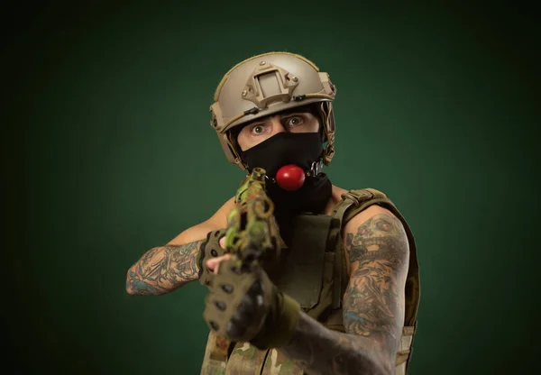 Ein männlicher Soldat in Militärkleidung, ein Helm mit einem Knebel im Mund drückt Emotionen aus, ein automatisches Gewehr in der Hand, Fotowitz — Stockfoto