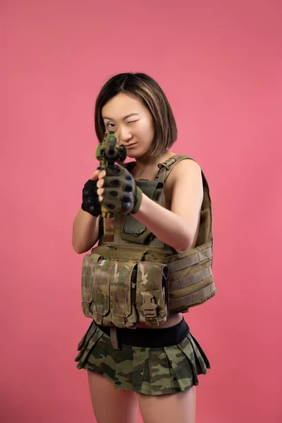 Sexy donna asiatica in abiti militari con un fucile automatico nelle sue mani su uno sfondo rosa — Foto Stock