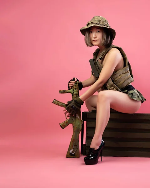 Askeri üniformalı, otomatik tüfekli seksi Asyalı kadın cephane sandığında oturuyor. — Stok fotoğraf
