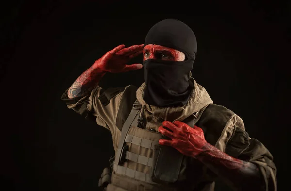 Foto de um homem com um uniforme militar e balaclava com um rosto sangrento e mãos até o cotovelo em sangue em um fundo preto — Fotografia de Stock