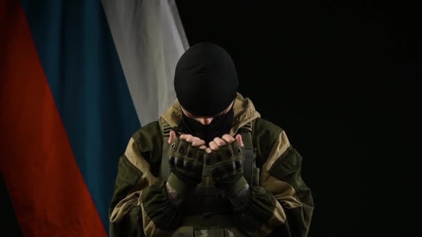 Un soldat russe sur le fond d'un drapeau russe avec un cagoule regrette et prie — Video