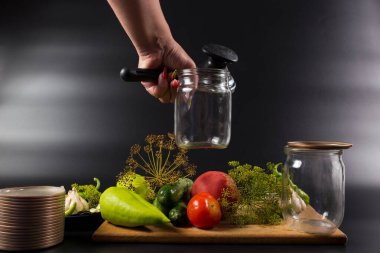 Ev yapımı sebze koruyucuları cam kavanozlarda otlar ve baharatlarla. Kadın, siyah arka planda cam kavanozlu konserve anahtarı tutuyor..