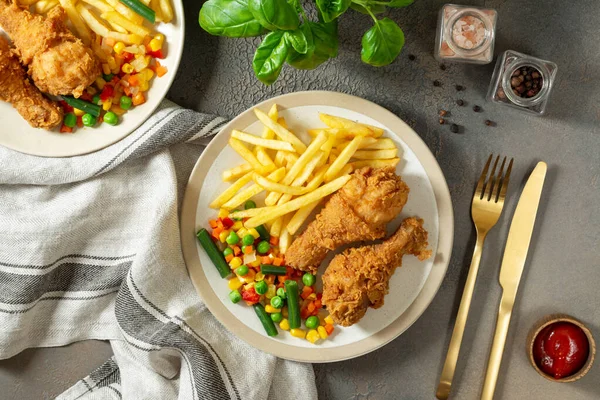 Fried crispy chicken legs in breadcrumbs with french fries and vegetables in a plate on a culinary background. Fast food from the drumstick top view