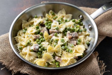 Yeşil bezelyeli Farfalle, hindi jambonu ve krema soslu parmesan mutfak masasındaki tavada. Koyu arka planda geleneksel İtalyan makarnası.