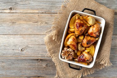 Açık gri ahşap mutfak arka planında, portakal, sarımsak ve soğanlı fırında pişirilmiş tavuk budu ve patates. Üst görünüm