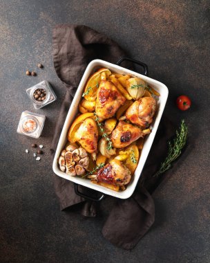 Fırında tavuk budu ve patates, portakal, sarımsak ve soğan ile birlikte karanlık bir mutfak arka planında pişirme kabında. Üst görünüm