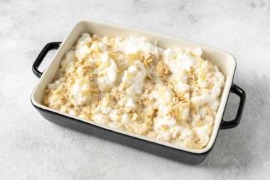 Açık gri mutfak arka planında fırın kabında çiğ puding ya da yulaflı turta. Mutfak masasının üzerinde fındıklı mısır gevreği var. Yemeği pişirmek için hazırlıyorum.