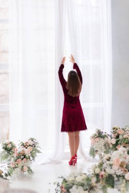 Elbiseli kadın pencerenin yanında duruyor ve perdeleri açıyor.