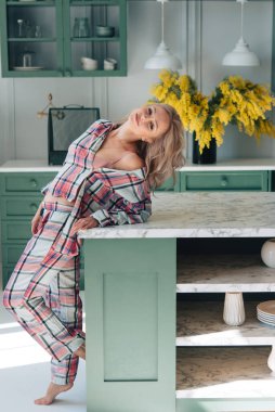 Evdeki mutfakta güzel, modern bir kadın. Ekose pijamalar. İç kısımda sarı çiçekler var. Tablo, yemekler, tasarım.
