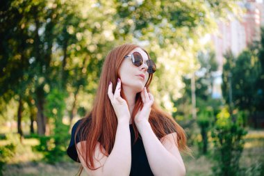 Siyah elbiseli, uzun saçlı güzel kızıl saçlı bir kadının portresi. Parktaki kadın. Yüzüne güneş gözlüğü..