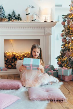 Güzel, şık, beyaz bir elbise giymiş küçük bir kız Noel ağacının yanında poz veriyor. Güzel dekore edilmiş Noel ağacı ve şömine, ev içi..