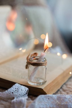 Eski açık bir kitabın arka planında cam bir kavanozda mum yakmak. Arka planda tatil ışıkları yanar.