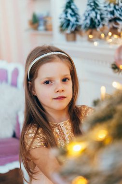 Güzel, şık, beyaz bir elbise giymiş küçük bir kız Noel ağacının yanında poz veriyor. Güzel dekore edilmiş Noel ağacı ve şömine, ev içi..