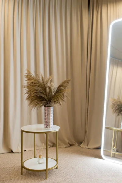 Soft home decor in gentle tones, a tall cylindrical vase with a bouquet of dried flowers against a background of beige curtains, on a round table. Interior.
