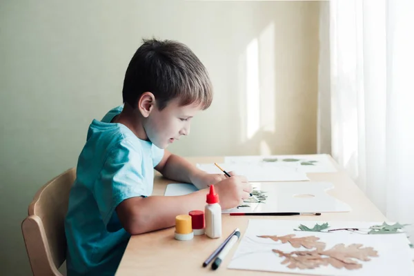 Anak bahagia duduk di meja dan membuat gambar dari daun kering — Stok Foto