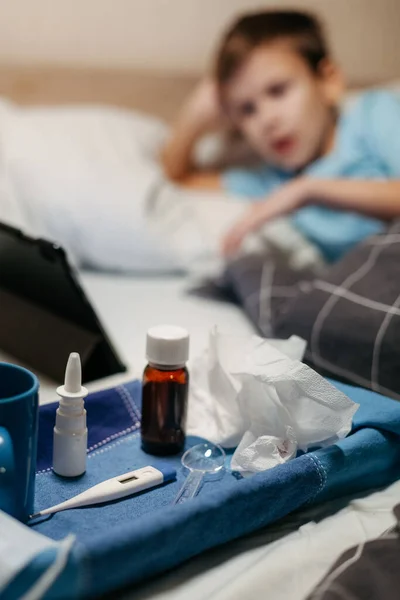 Frasco com xarope antipirético, spray nasal e termômetro em guardanapo. Menino na cama em primeiro plano fora de foco — Fotografia de Stock