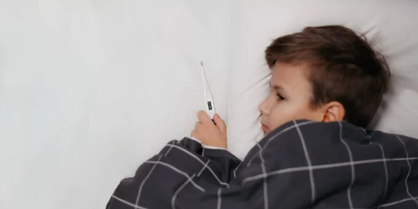 Criança doente deitada na cama e segurando termômetro. DoF pequeno, foco no termômetro — Fotografia de Stock