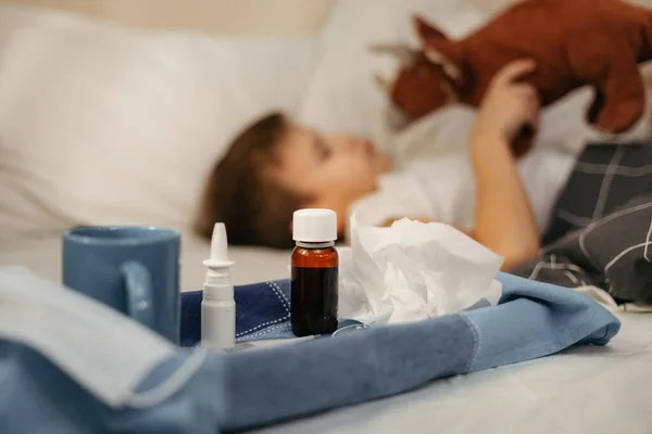 Botol dengan sirup antipiretik, semprotan hidung dan termometer pada serbet. Anak kecil di tempat tidur di latar depan keluar dari fokus — Stok Foto