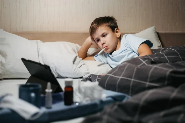 Anak sedih berbaring di tempat tidur dengan termometer di mulutnya dan melihat tablet — Stok Foto