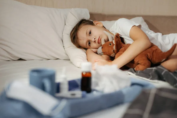 Mały chłopiec leżący w łóżku z zabawką. Leki i termometr na pierwszym planie nieostre. — Zdjęcie stockowe