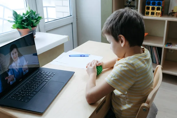 Niño de 8 años sentarse en el escritorio con el ordenador portátil y hacer ejercicio con bola de masaje Imagen de archivo