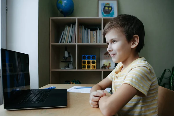 8 살 소년은 책상 옆에 노트북을 놓고 앉아서 온라인 수업중에 과제를 작성 합니다. — 스톡 사진