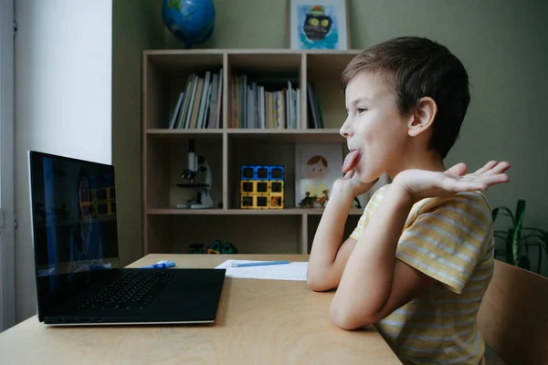 8 tahun anak laki-laki tua duduk dengan meja dengan laptop dan mencuat lidahnya dan main-main — Stok Foto