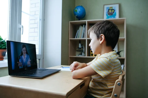 8 jaar oud jongen zitten door bureau met laptop en doen schrijven taak tijdens online les Rechtenvrije Stockfoto's