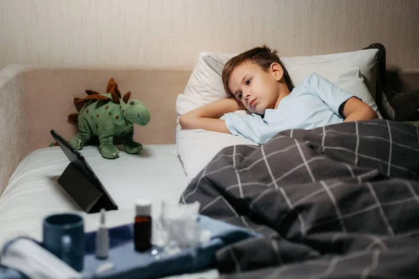Anak sakit yang sangat menyedihkan berbaring di tempat tidur dengan mainan dan tablet — Stok Foto