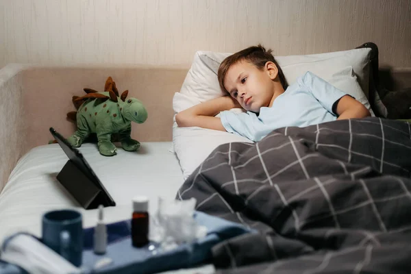 Zeer verdrietig ziek kind liggend in bed met speelgoed en tablet Stockfoto