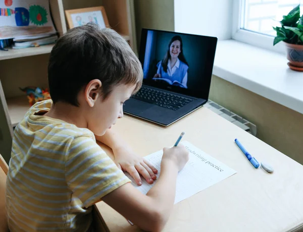 8 tahun anak laki-laki tua duduk dengan meja dengan laptop dan melakukan tugas menulis selama pelajaran online — Stok Foto