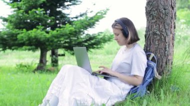 Güzel genç kız ağacın yakınındaki parkta ders çalışıyor. Genç bir kadın çalışmak için dizüstü bilgisayar kullanıyor. Küçük yakınlaştırma videosu