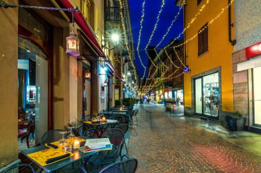 Kuzey İtalya, Piedmont 'taki eski Alba kasabasının Noel ışıklarıyla aydınlatılmış dar kaldırım taşı caddesinde mumlarla dolu restoran masaları..